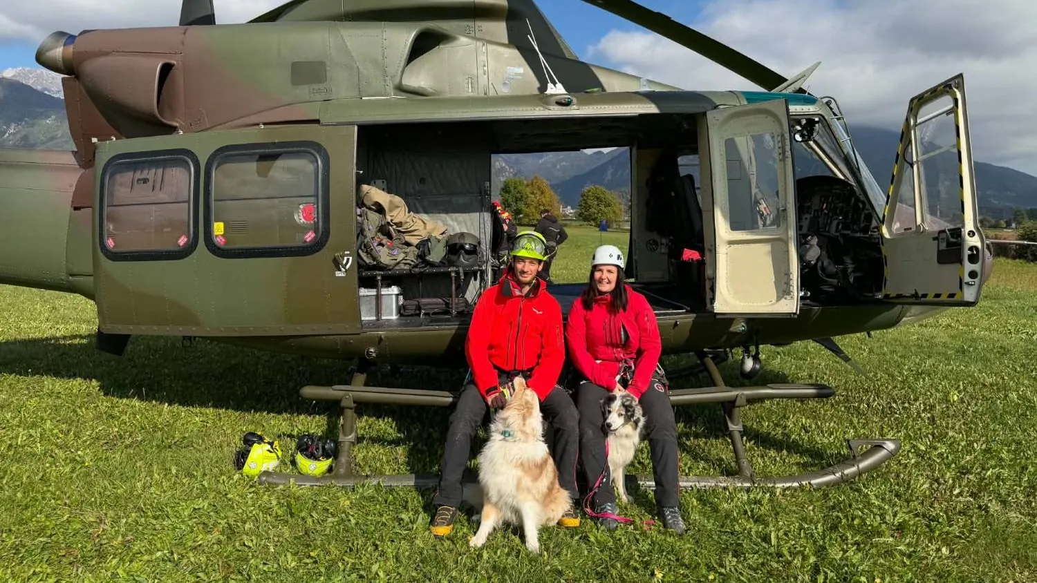 Trije vodniki s psi z nagobčniki na travniku, v ozadju helikopter.