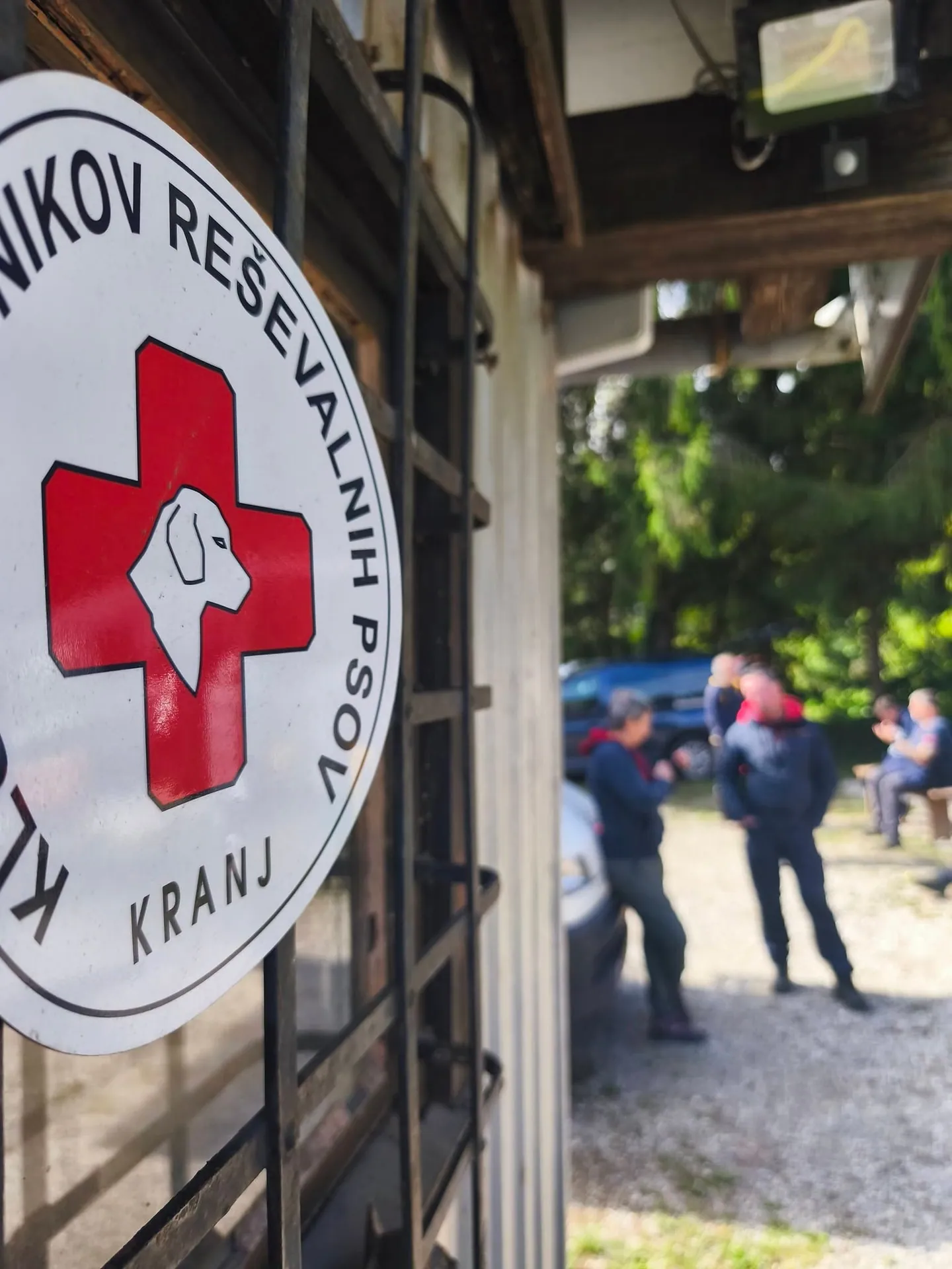 Znak Kluba vodnikov reševalnih psov Kranj na klubskem objektu, v ozadju udeleženci.